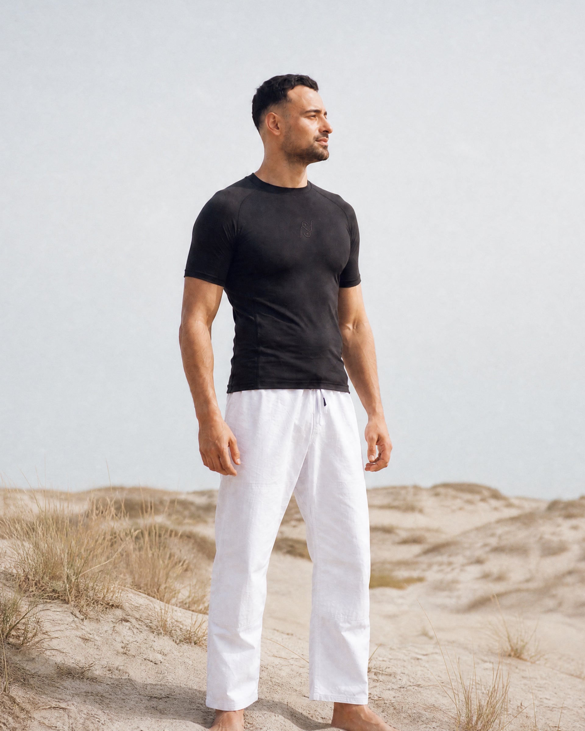 Man on beach dunes wearing black V1 short sleeve rashguard with white gi pants, front
